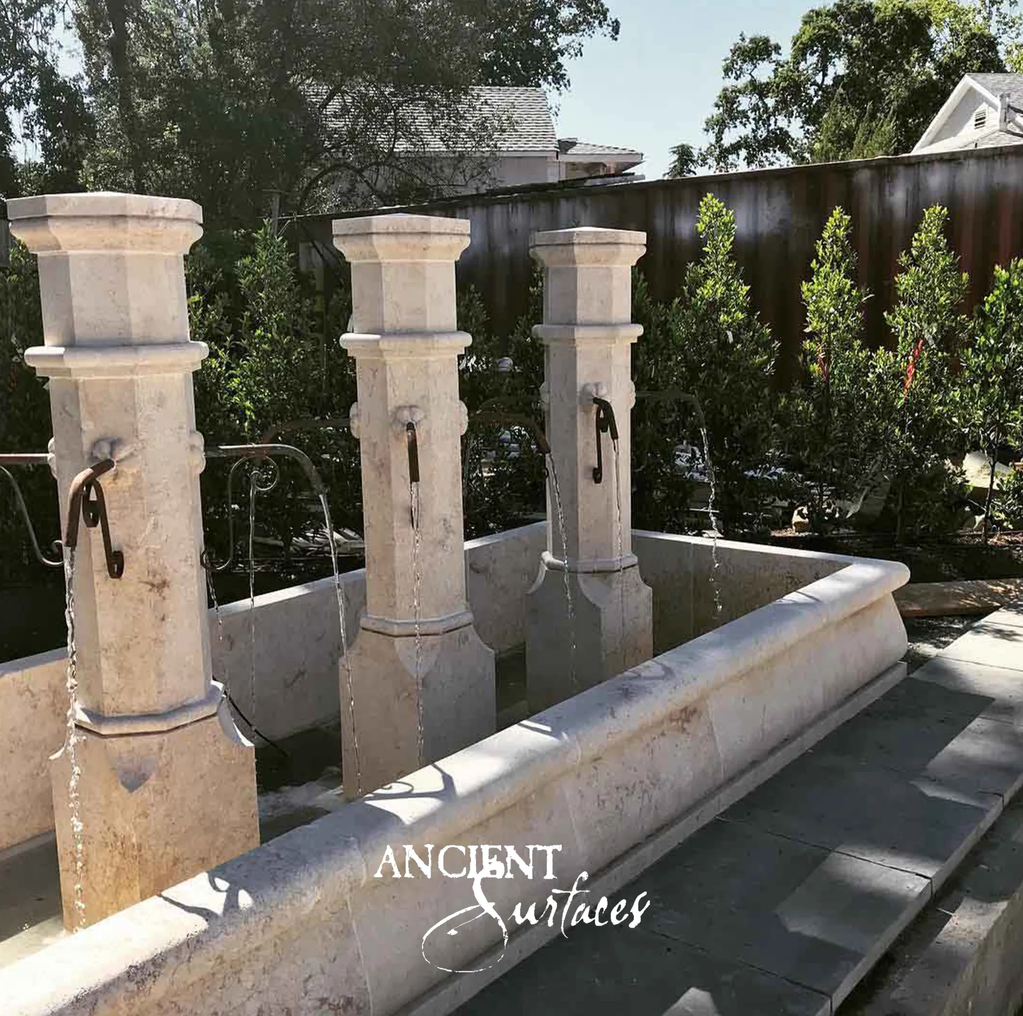 Three reclaimed limestone pillar fountains from Ancient Surfaces pouring into a long stone trough in a sunlit garden